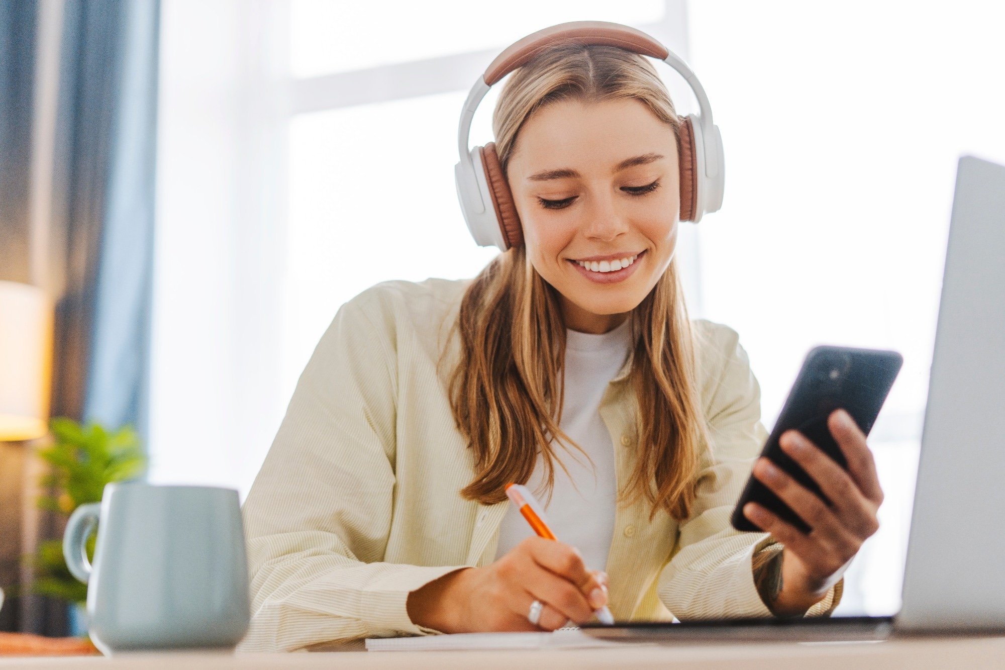 Smiling woman takes notes at her home office, wearing headphones listening music at home using mobile phone remote work.