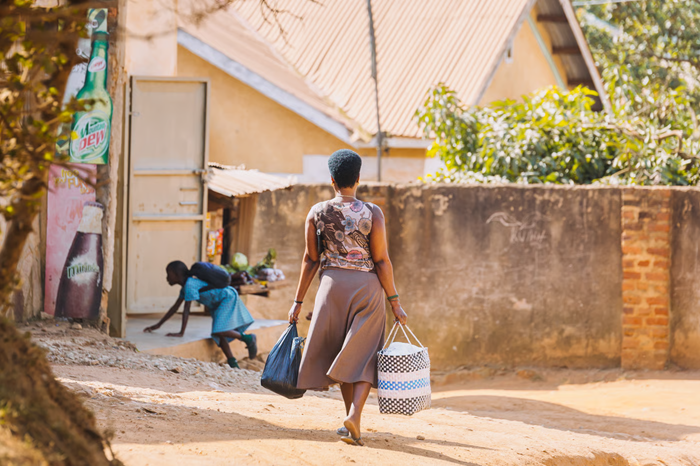 A woman with short black hair carrying shopping bags in both hands, walking through the landscape of a hot, dry country.