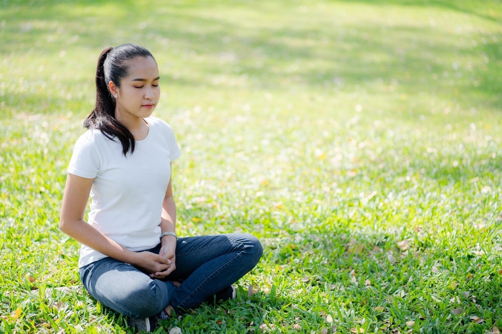 A young woman meditating peacefully outdoors on a sunny day, sitting cross-legged on green grass in a relaxed posture.