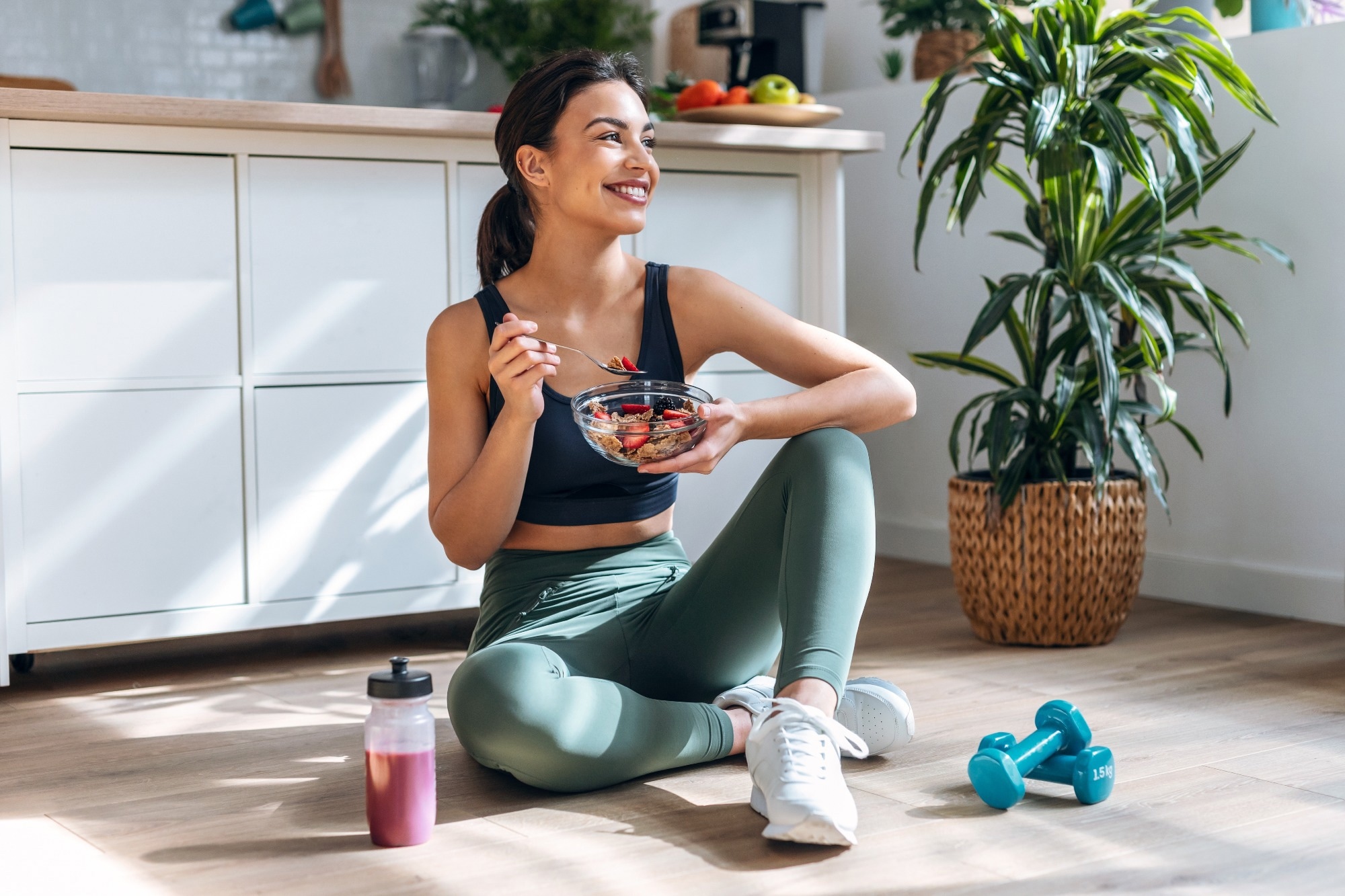 Shot of athletic woman eating a healthy bowl of muesli with fruit sitting on floor in the kitchen at home.