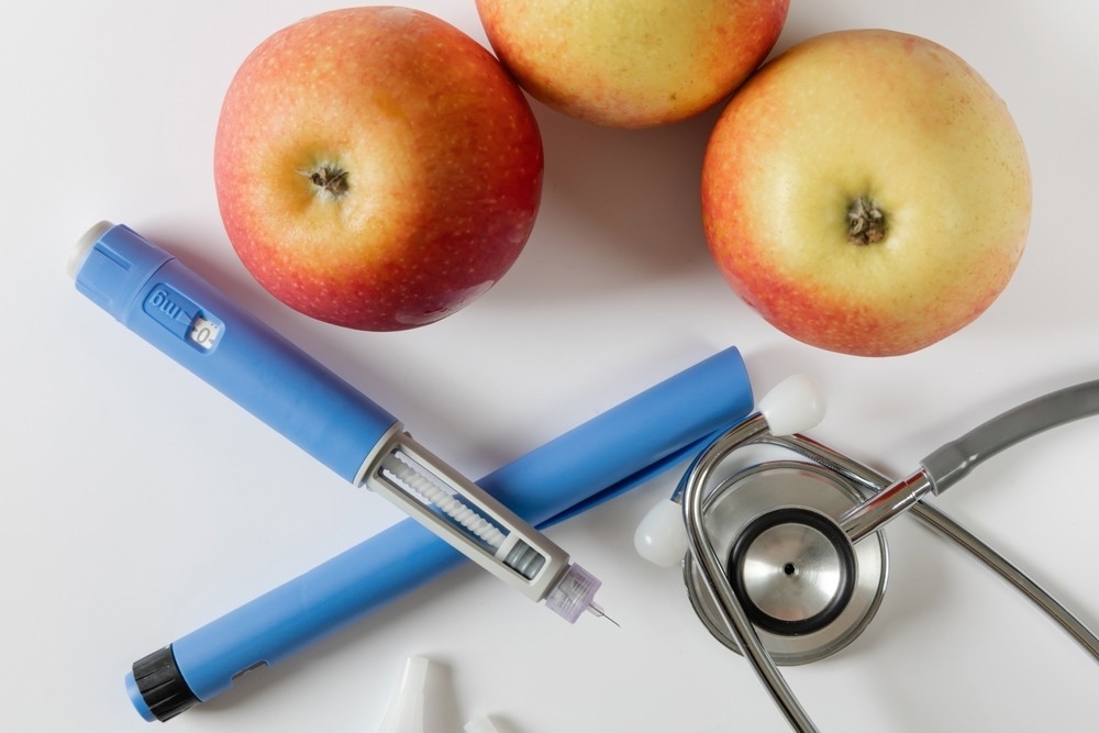 A close-up of three apples, two blue insulin pens, and a stethoscope arranged on a white background, symbolizing diabetes management, healthy eating, and medical care.