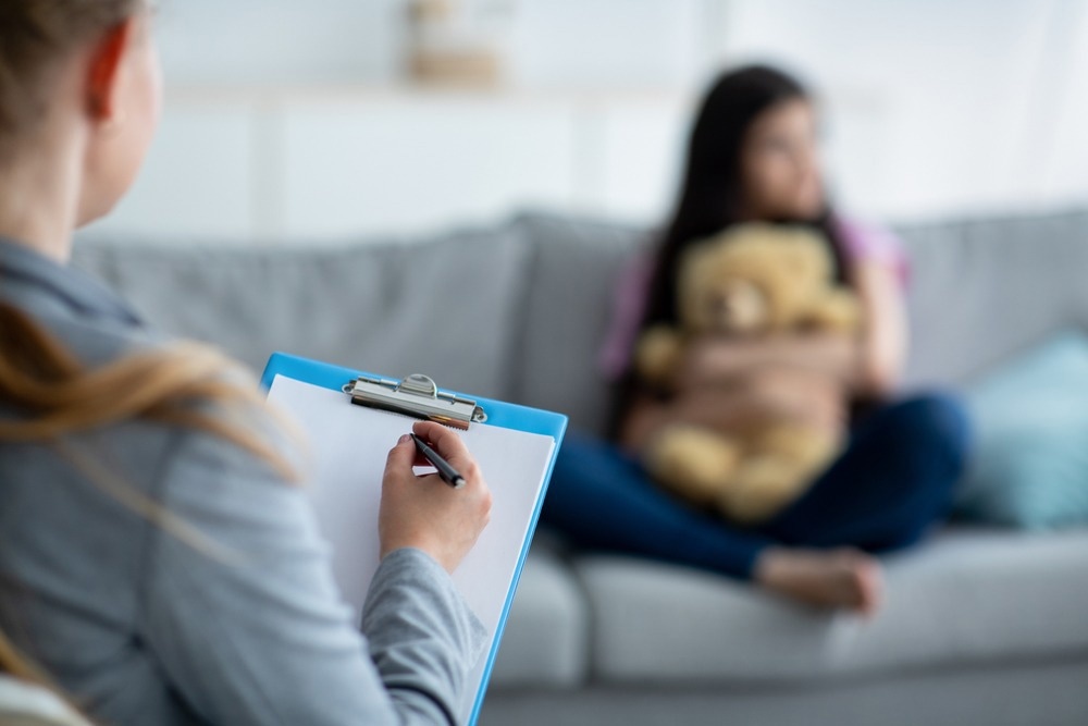A blurred young girl sitting on a couch hugging a teddy bear, while a mental health professional in the foreground takes notes on a clipboard. The setting appears to be a therapy session in a well-lit room.