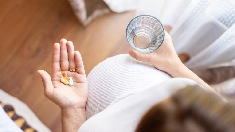 A pregnant woman in a white top holds a glass of water in one hand and a few prenatal vitamins in the other. The image is taken from a top-down perspective, highlighting her baby bump and the supplements she is about to take.