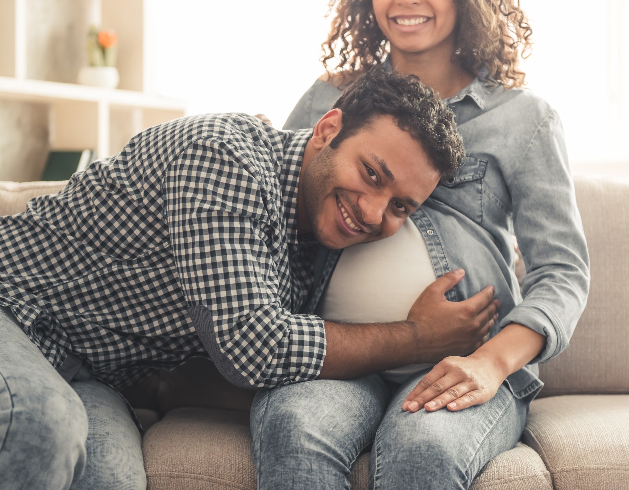 Handsome Afro American man and his beautiful pregnant wife are sitting on couch and smiling while spending time together at home.