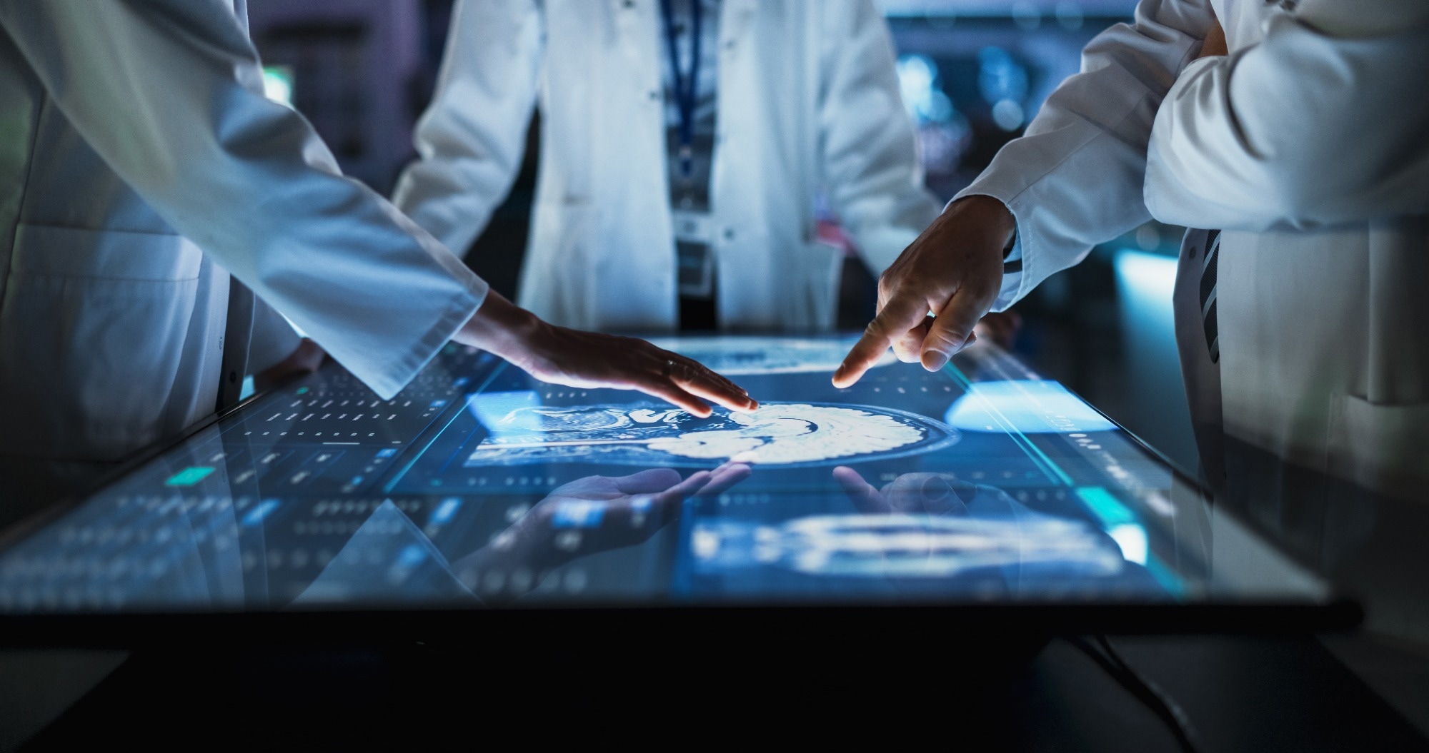 Focus On Hand Gestures Of Diverse Colleagues Gathered Around Interactive Touch Screen Table With MRI Scans Of Brain On Display.