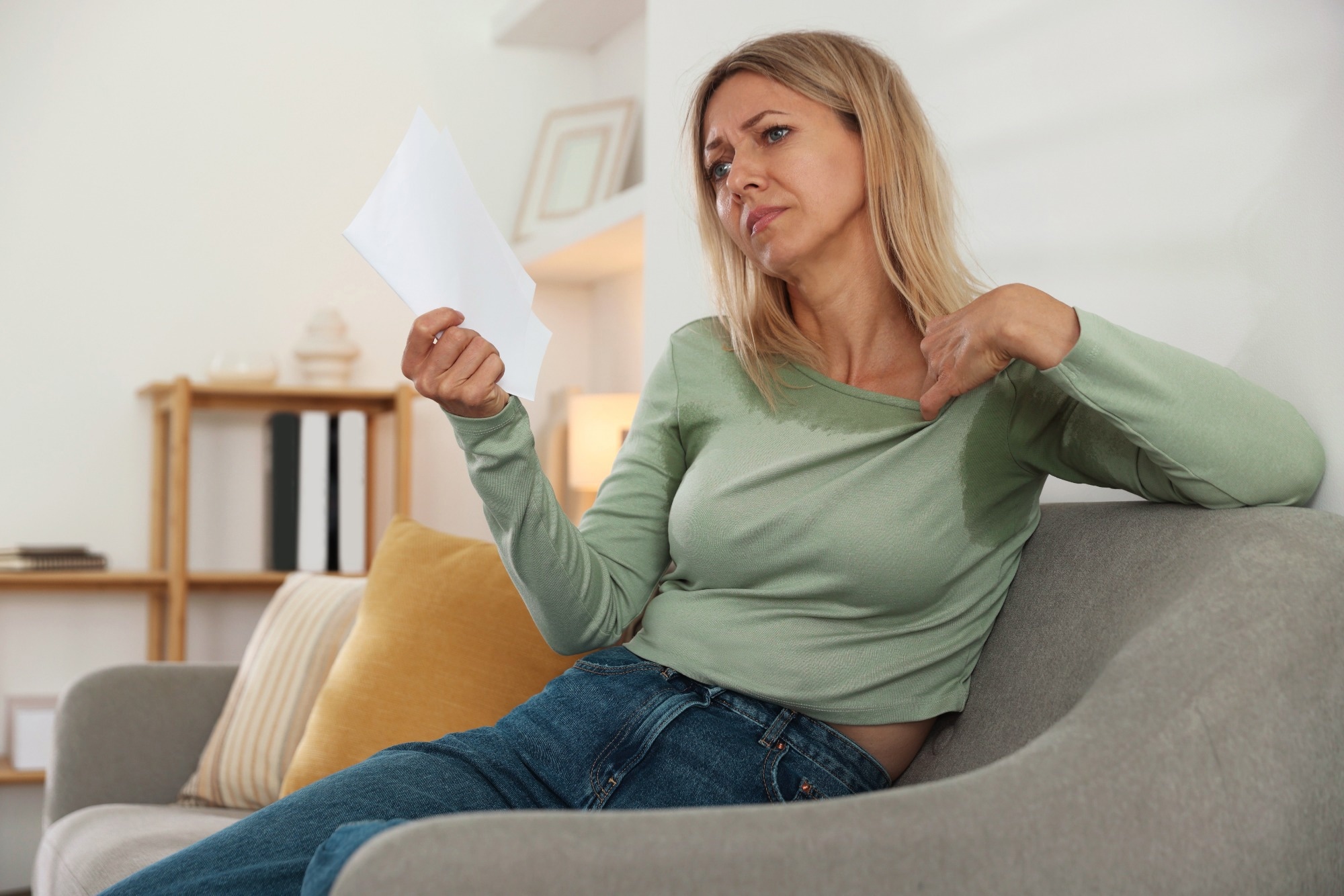 Woman waving paper sheet to cool herself during hot flash on sofa at home