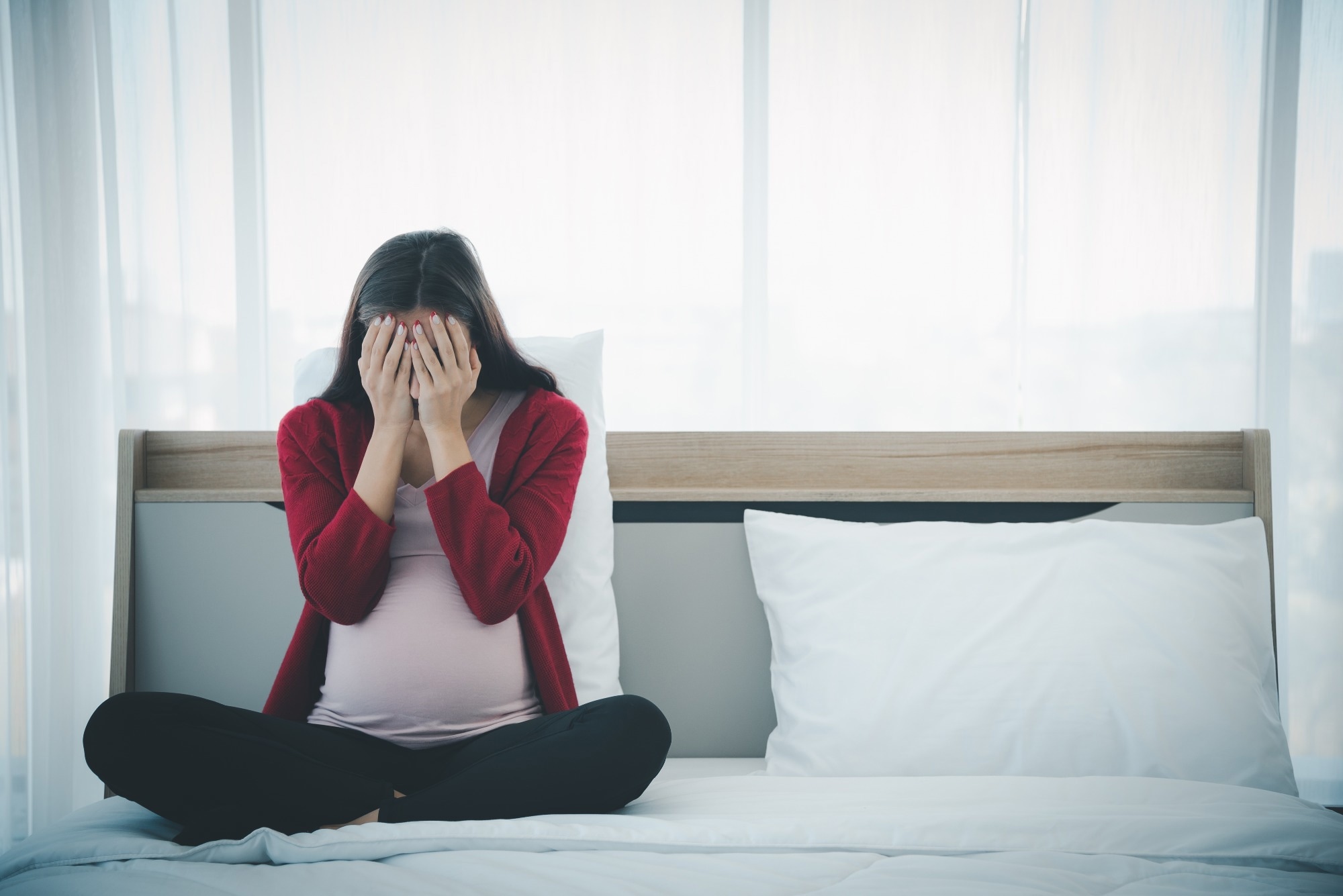 Pregnant woman in casual home outfit sitting on bed and feeling upset and sad