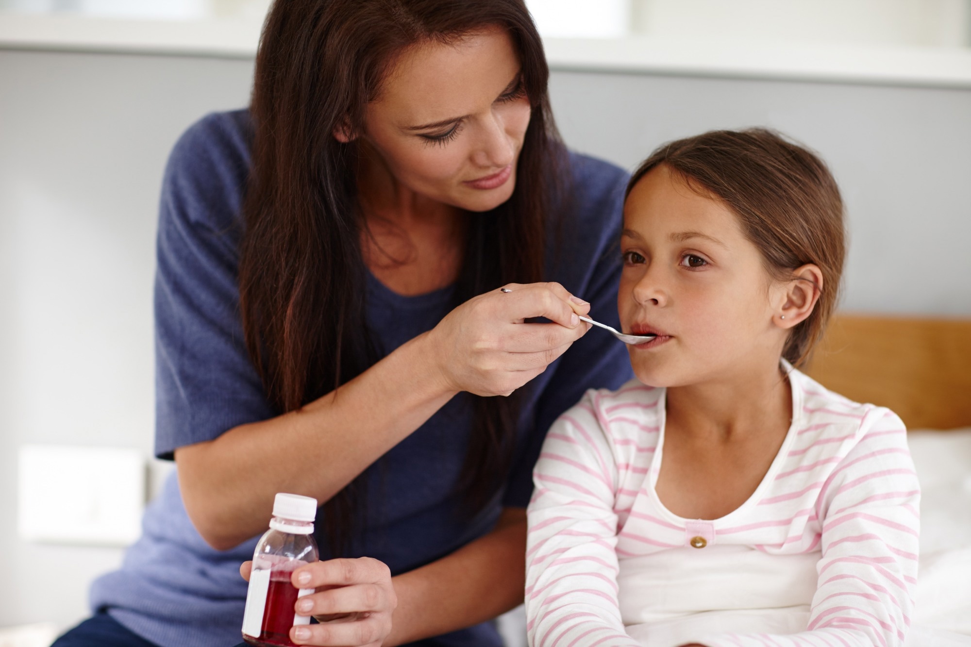 Mother holding a bottle of medicine. Mother giving sick young daughter a spoon of antibiotic medicine. Daughter is in bed.